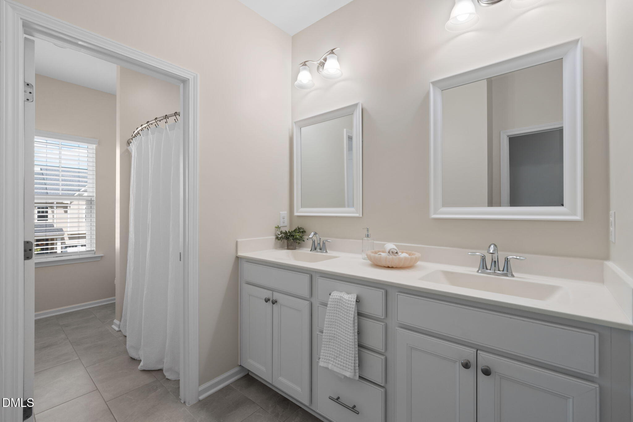 Bathroom with a double-sink vanity, gray cabinets, framed mirrors, and a curtained shower beside a window.