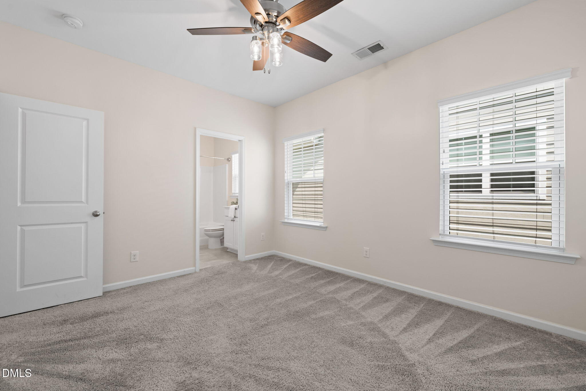 Empty carpeted bedroom with a ceiling fan, two blinded windows, and an open bathroom doorway.