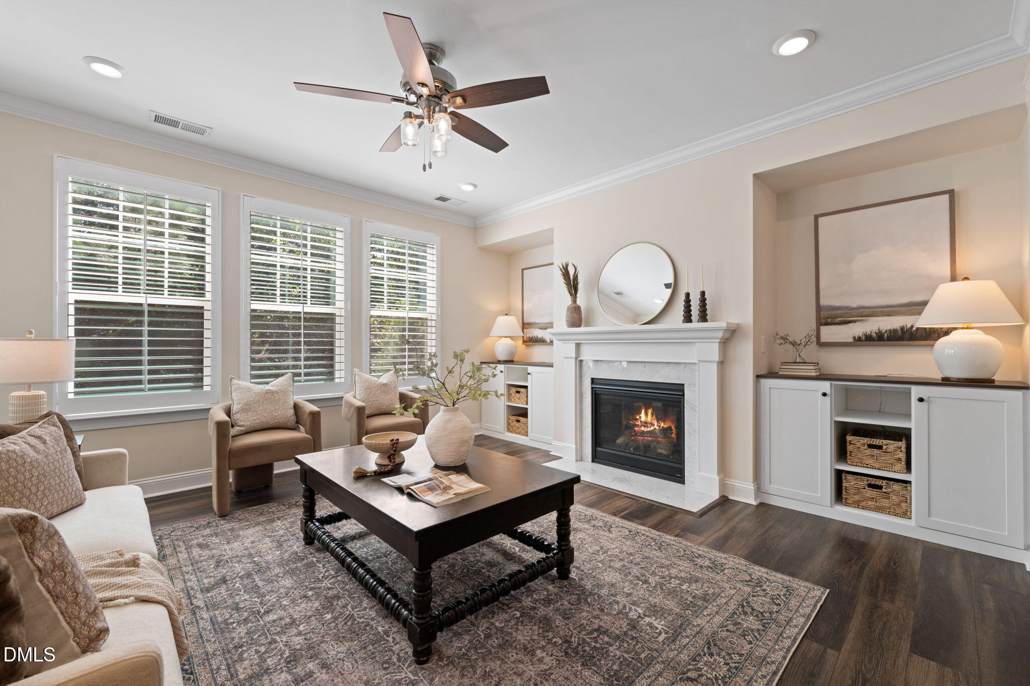Staged living room with a lit fireplace, plantation shutters, and white built-in cabinetry.