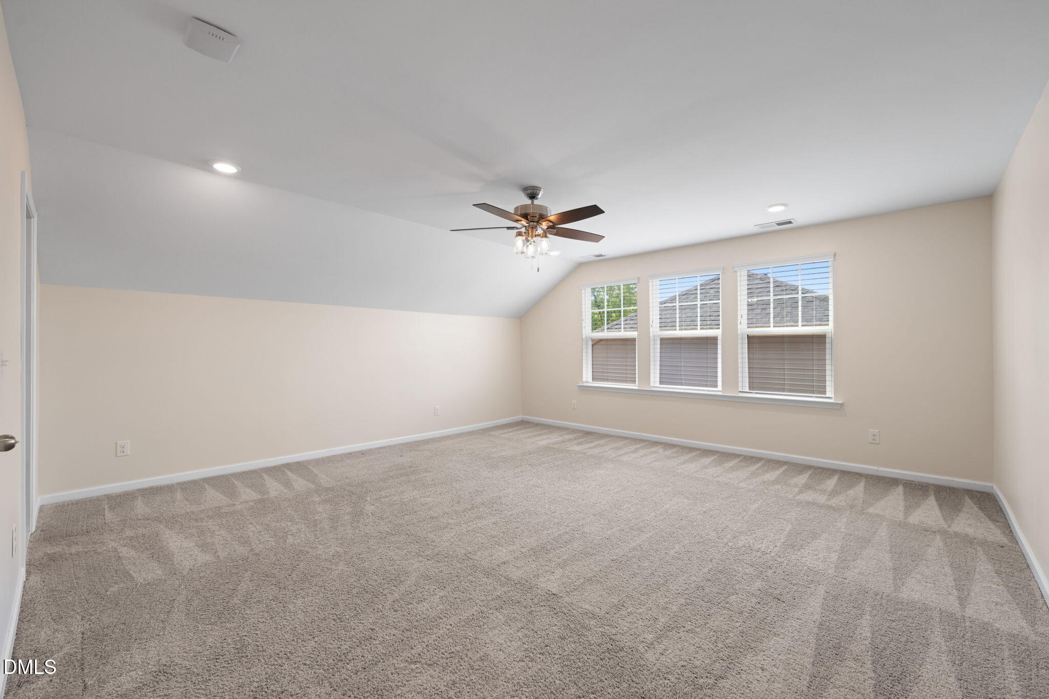 Empty carpeted room with a sloped ceiling, ceiling fan, and three windows with blinds.