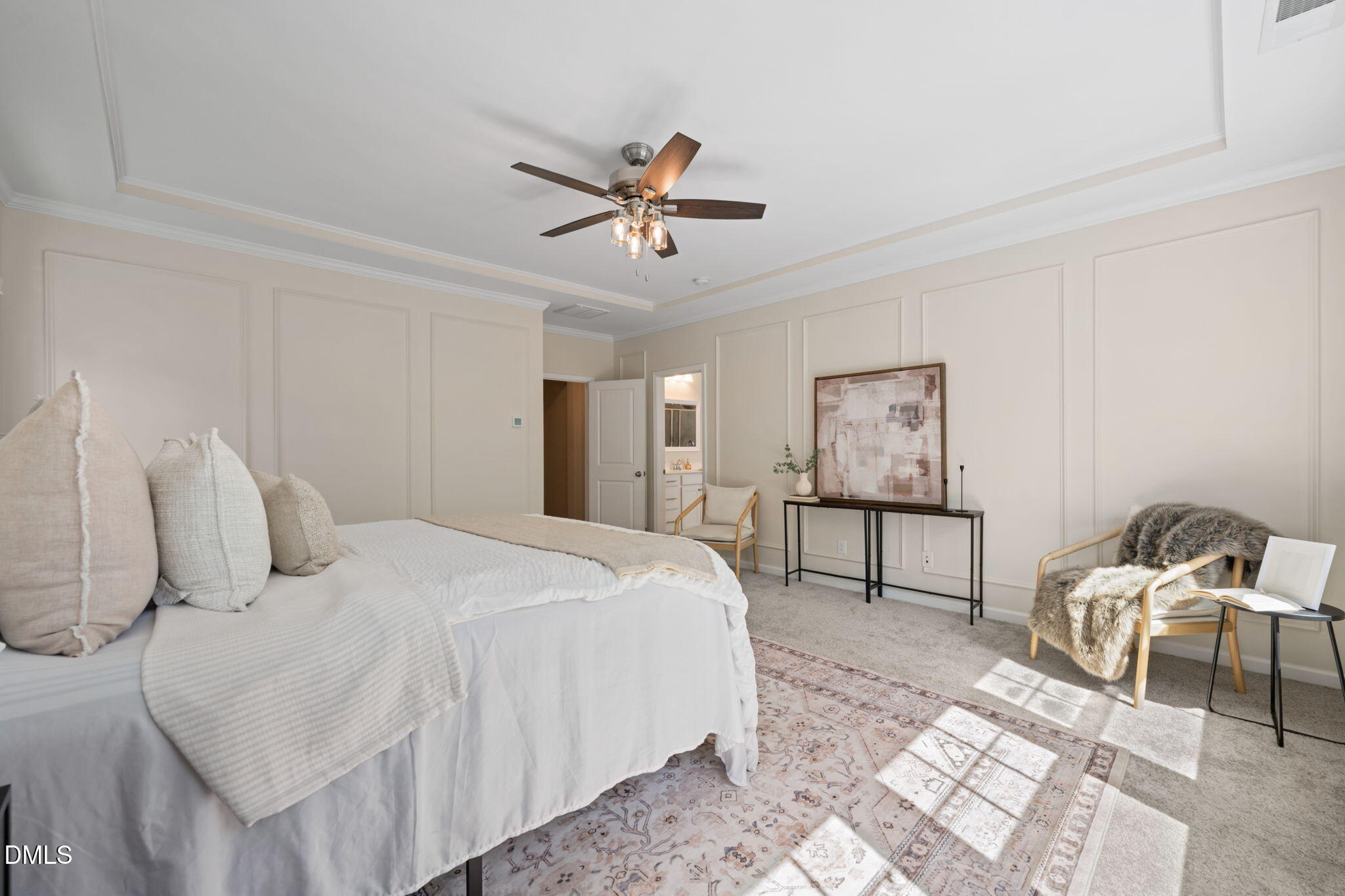 Staged bedroom with a white bed, paneled walls, ceiling fan, and sunlit sitting area.
