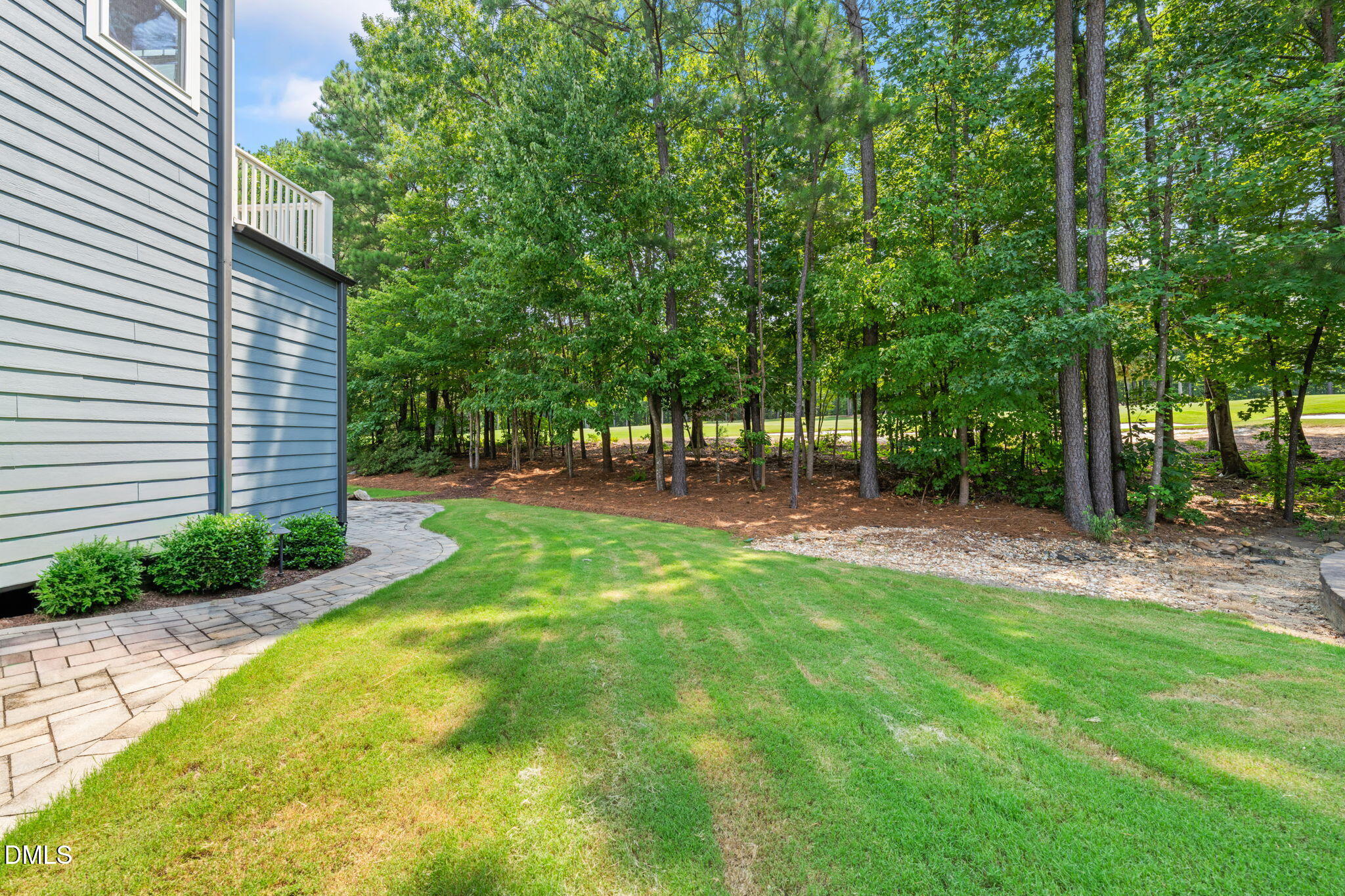 Backyard lawn and curved stone walkway beside a blue-sided house, bordered by mature trees.