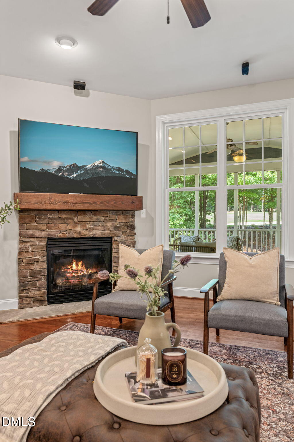 Staged living room with a stone fireplace, two chairs, and a large window overlooking trees.