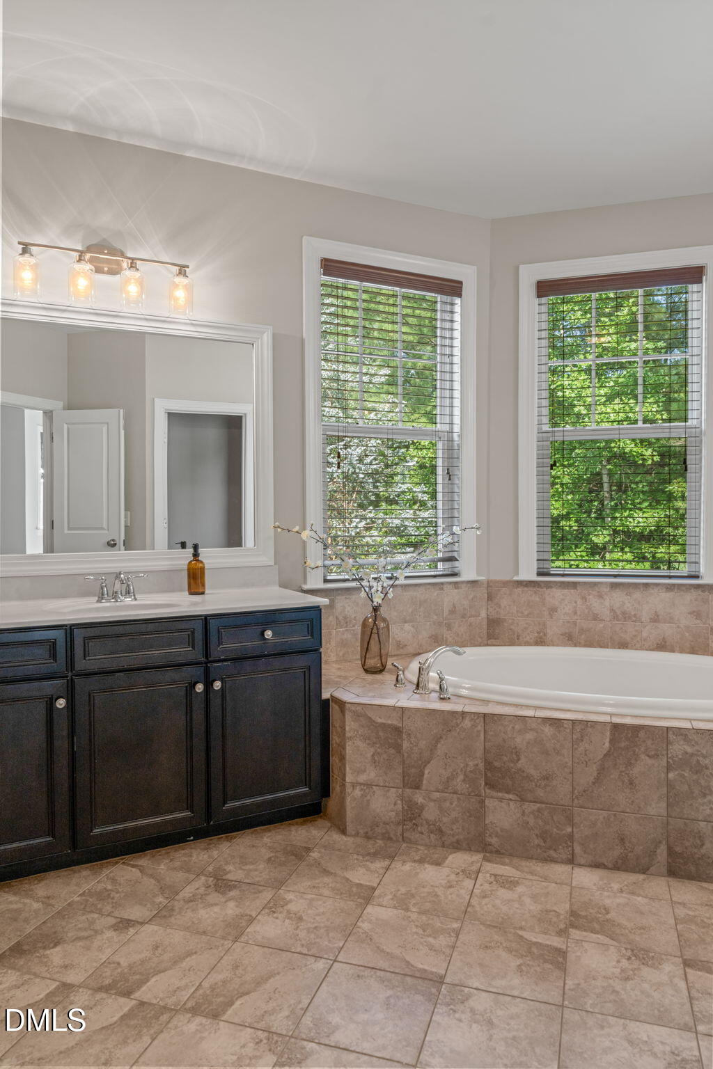 Bathroom with a dark blue vanity and tiled soaking tub beneath two leafy-window views.