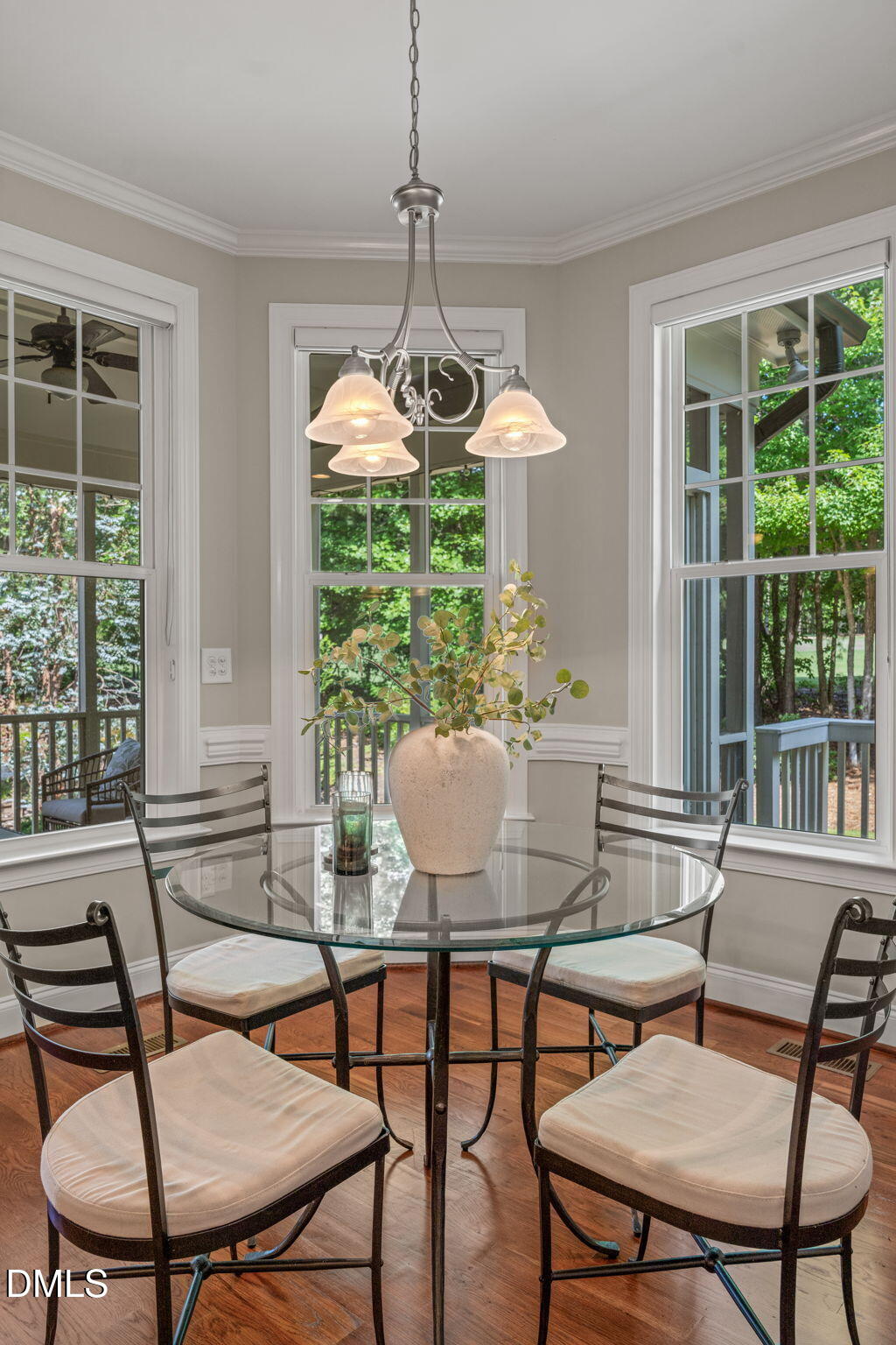 Dining room with a glass table, cushioned chairs, chandelier, and large windows overlooking trees.