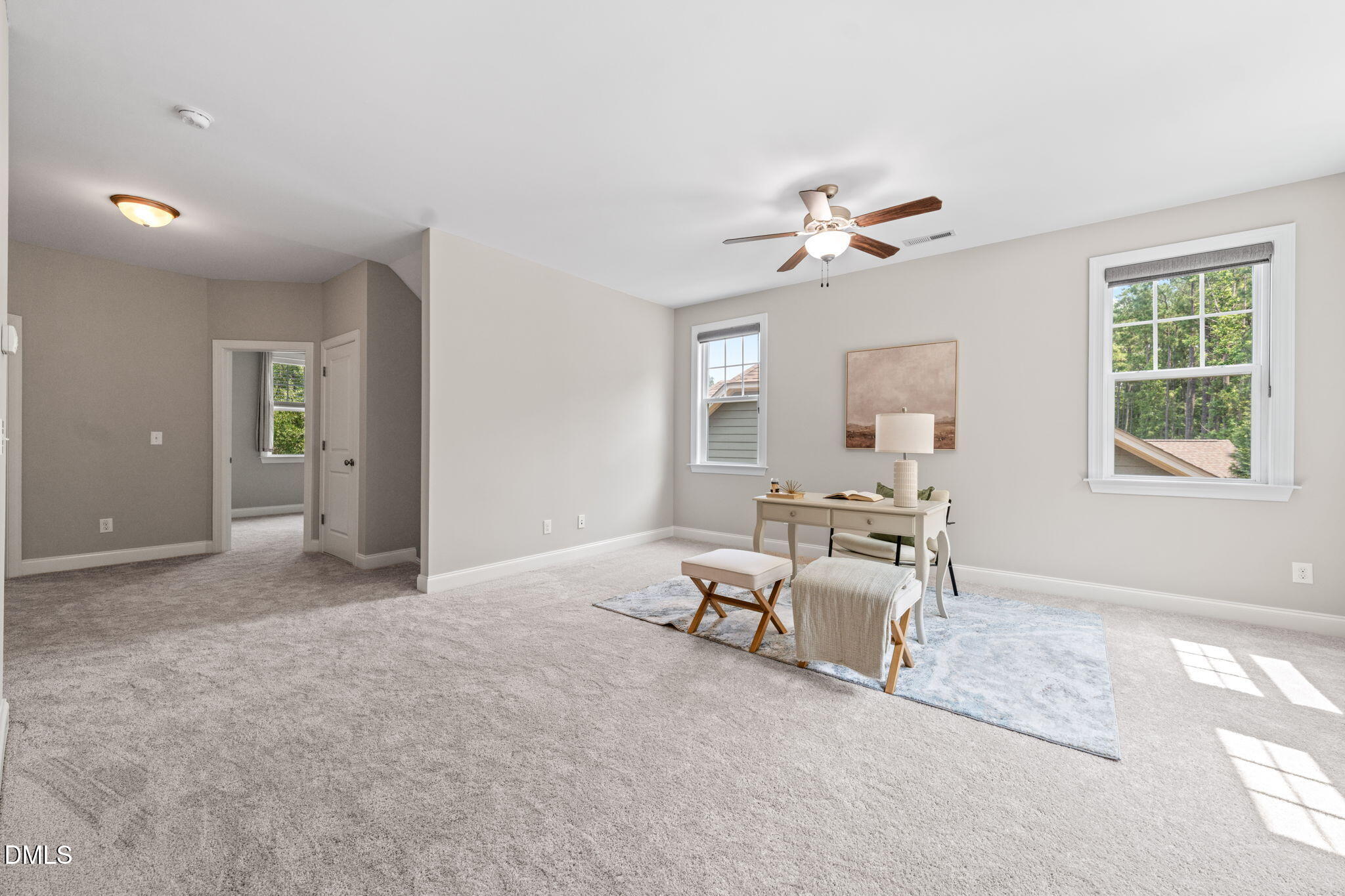 Carpeted bedroom staged as an office, with a desk, ceiling fan, and windows overlooking trees.