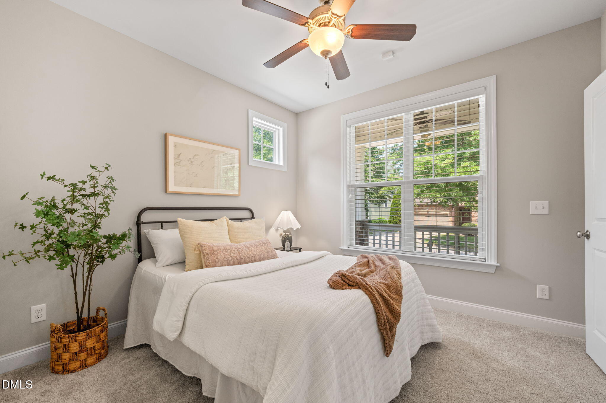 Staged bedroom with a white bed, ceiling fan, and large window overlooking greenery.