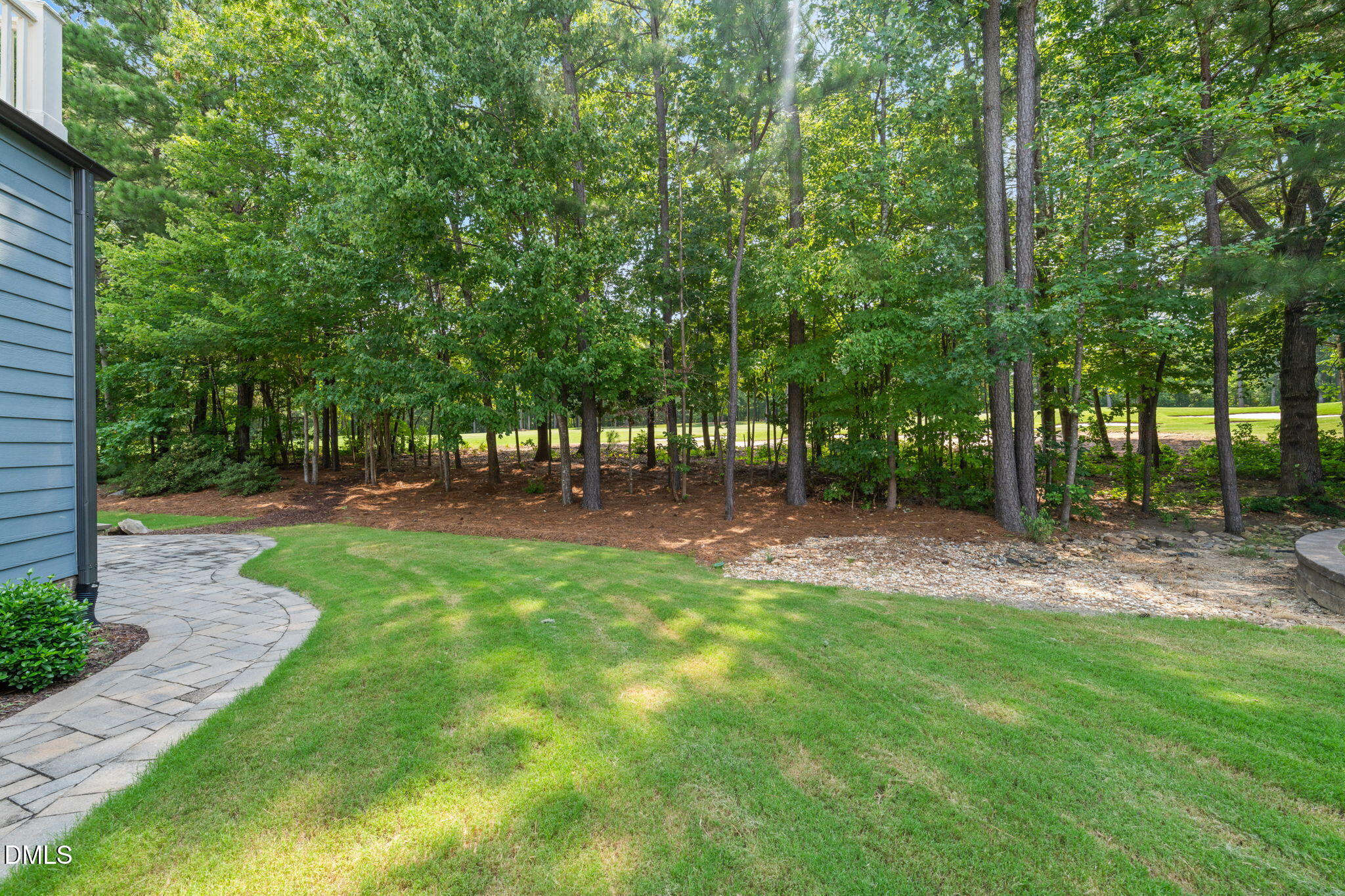 Backyard lawn and curved stone walkway beside a blue-sided house, with mature trees along the back.