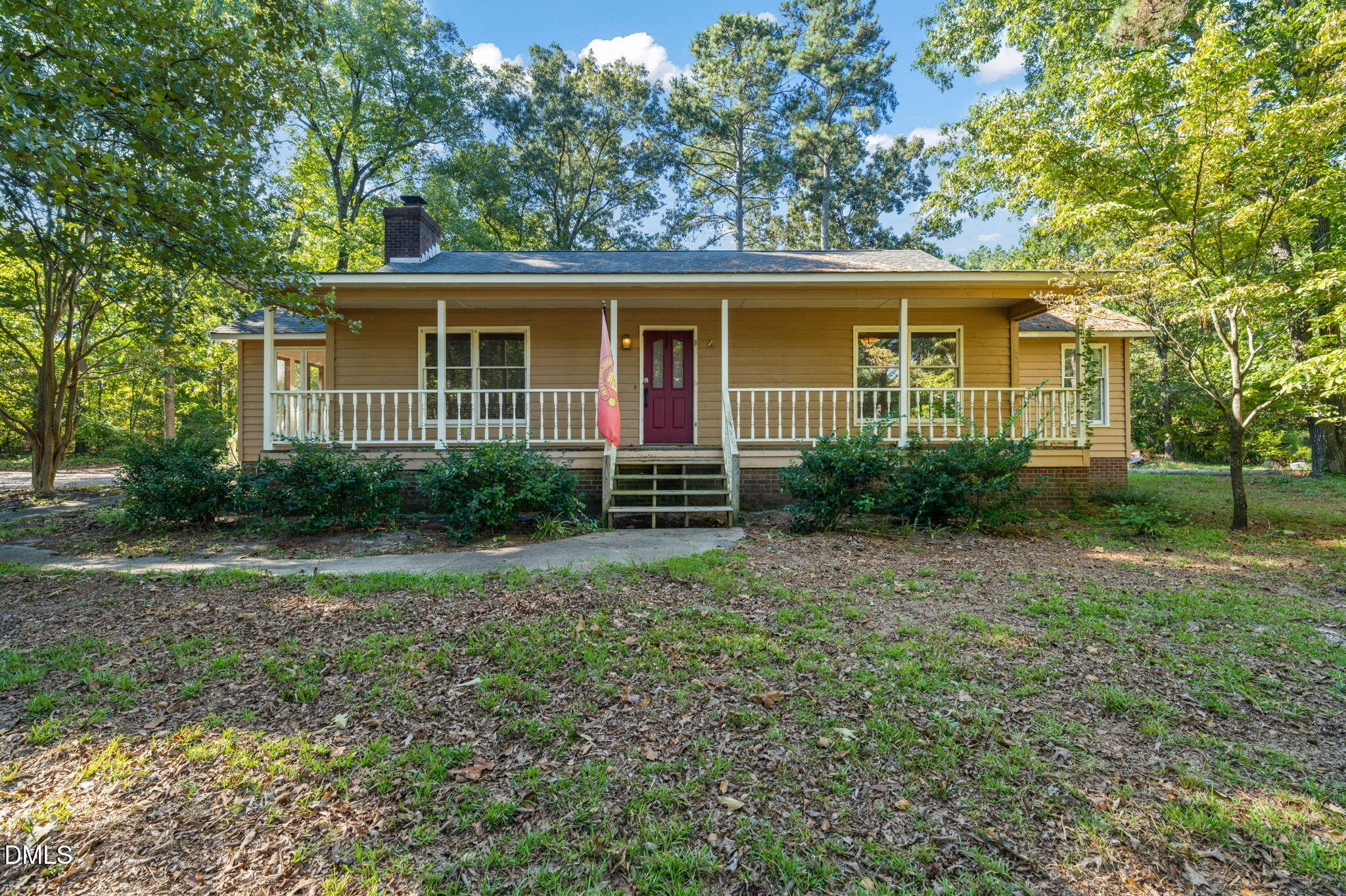 Tan single-story home with a long covered porch and burgundy front door, surrounded by mature trees.