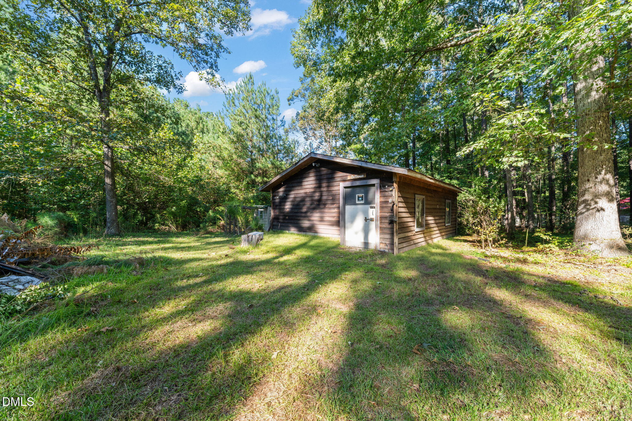 Wood-sided outhouse in a grassy, tree-lined clearing, with a white door and two windows.