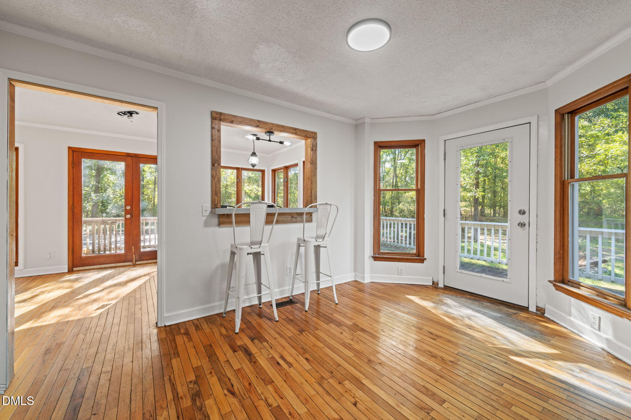 Dining area with polished hardwood floors, a pass-through counter with two stools, and windows overlooking trees.