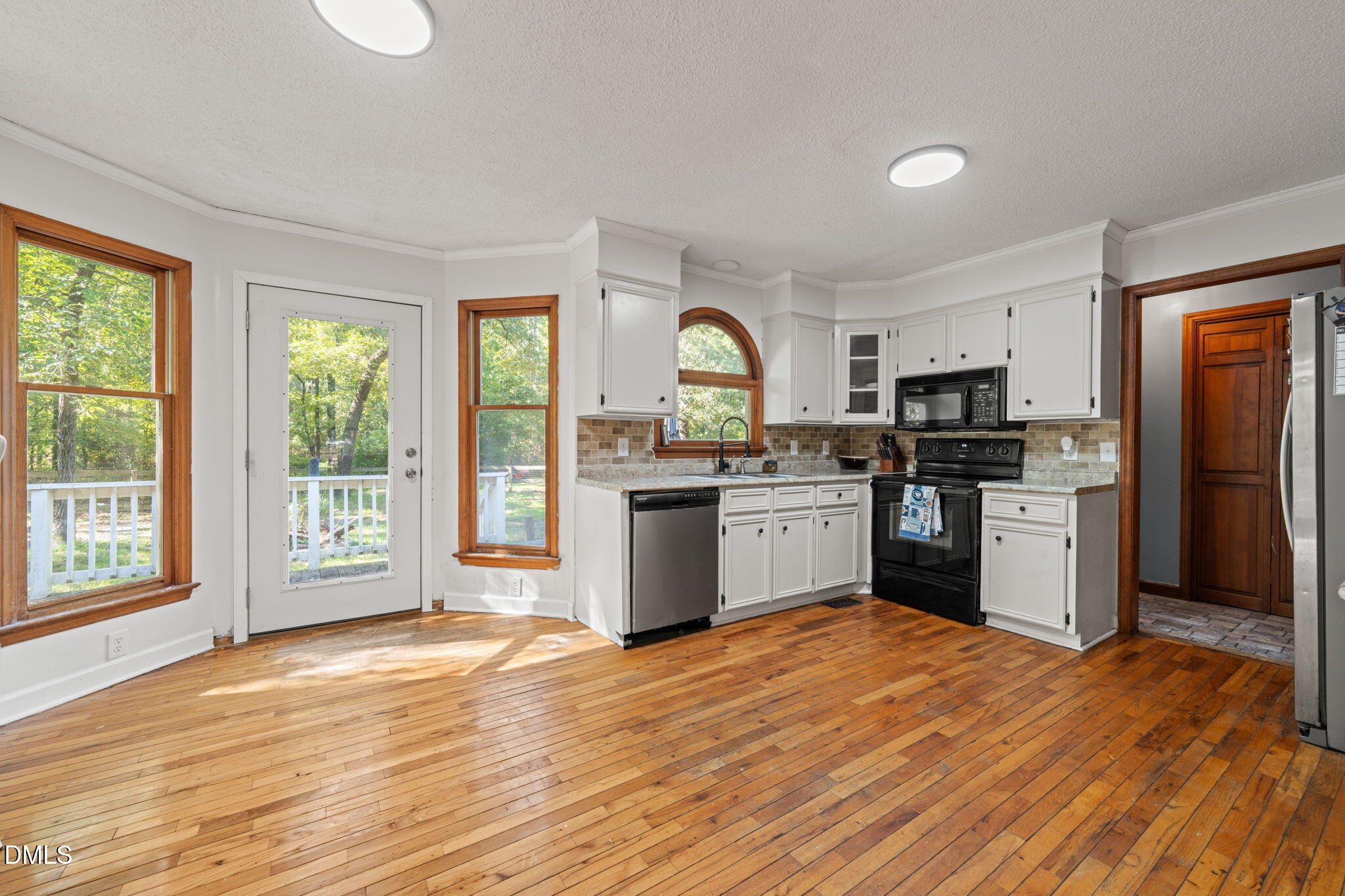 Kitchen with white cabinetry, hardwood floors, and wood-trimmed windows overlooking a leafy yard.