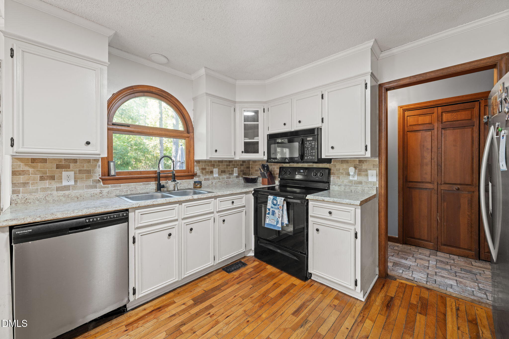 Kitchen with white cabinets, an arched wood-trimmed window, hardwood floors, and black appliances.
