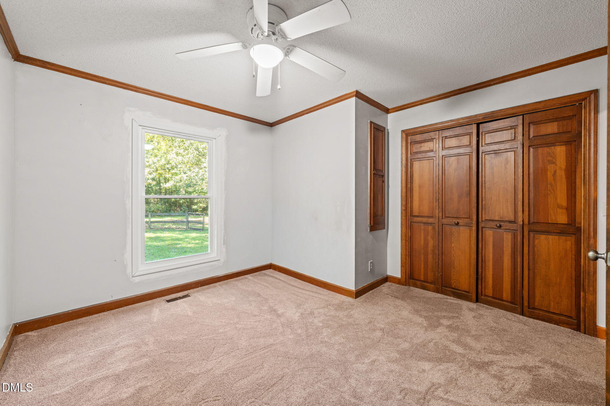 Empty bedroom with built-in wood closet doors, beige carpet, and a window overlooking a fenced lawn.