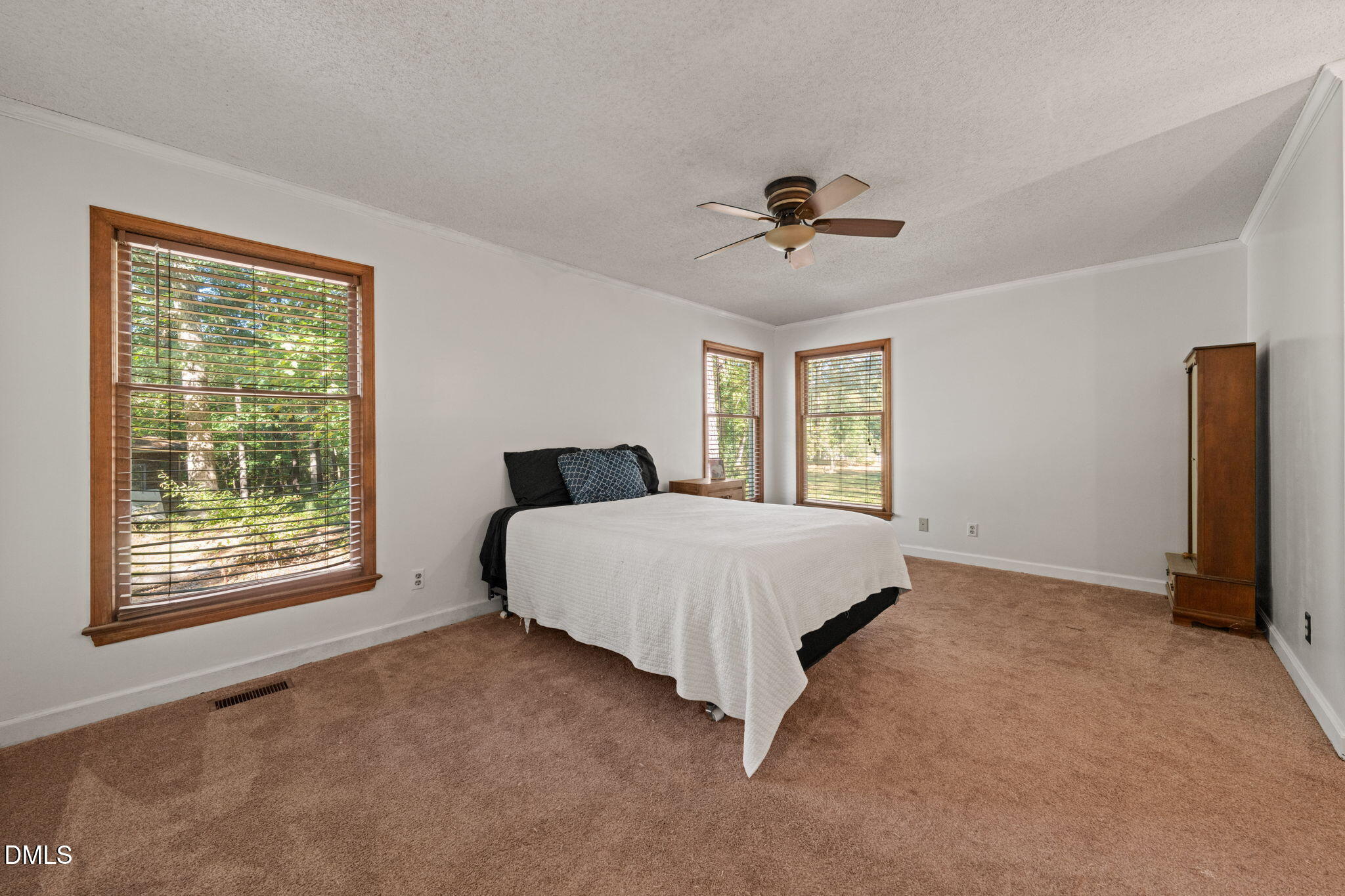 Bedroom with a white-covered bed, brown carpet, and three wood-trimmed windows with blinds.