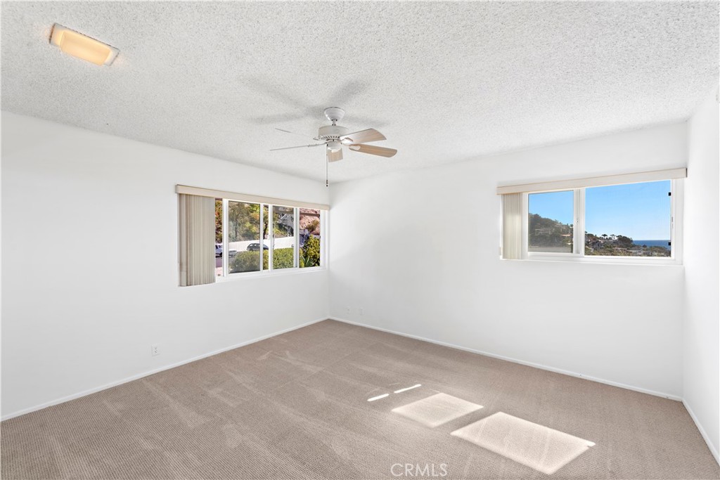 Empty carpeted bedroom with a ceiling fan and windows showing coastal hills and a glimpse of the ocean.