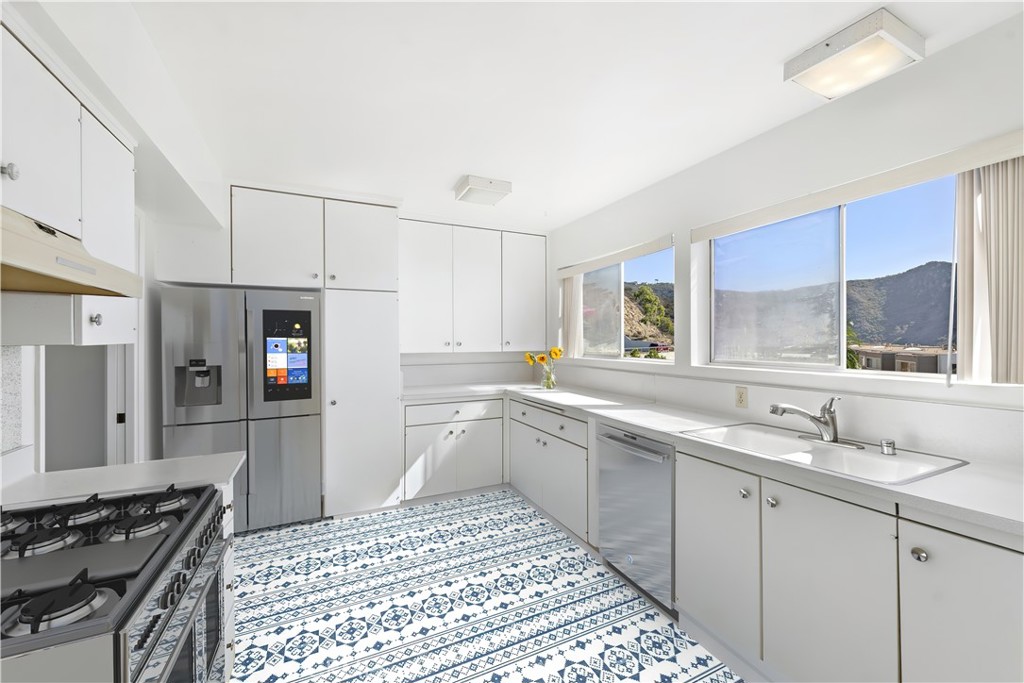 Bright white kitchen with stainless appliances, patterned blue-and-white tile floor, and hillside views through large windows.