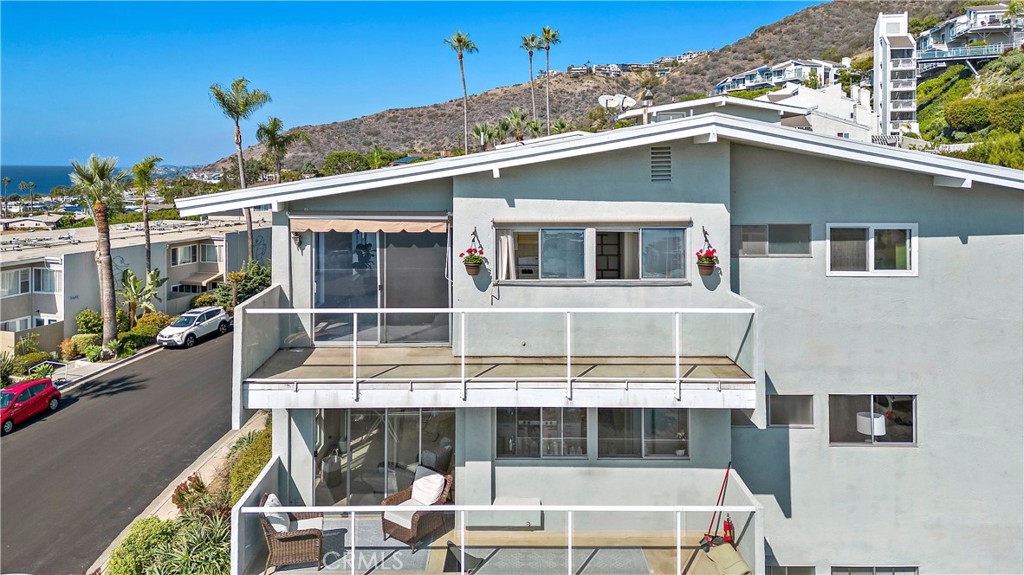 Coastal condominium exterior with glass-railed balconies, hillside homes, palm trees, and a glimpse of ocean.