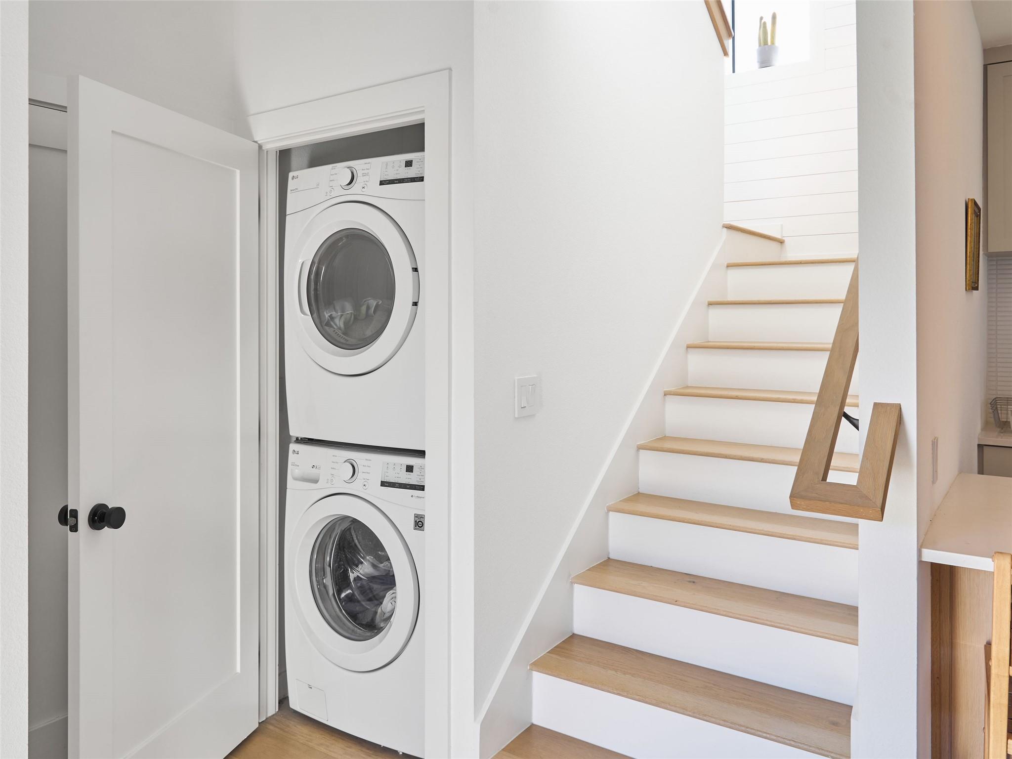 Stacked washer and dryer beside a staircase with light wood treads and a simple wood handrail.