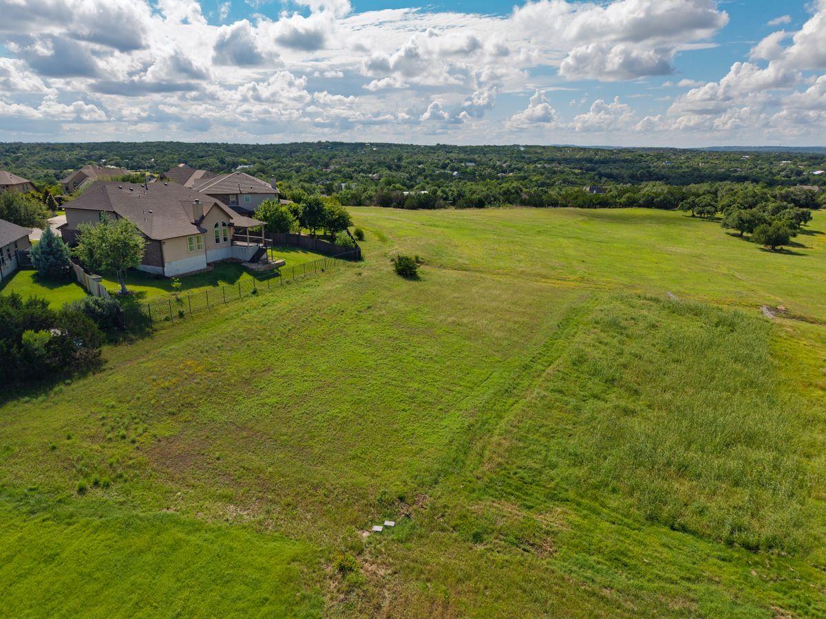 Aerial view of a broad grassy lot beside a home, with wooded landscape beyond.