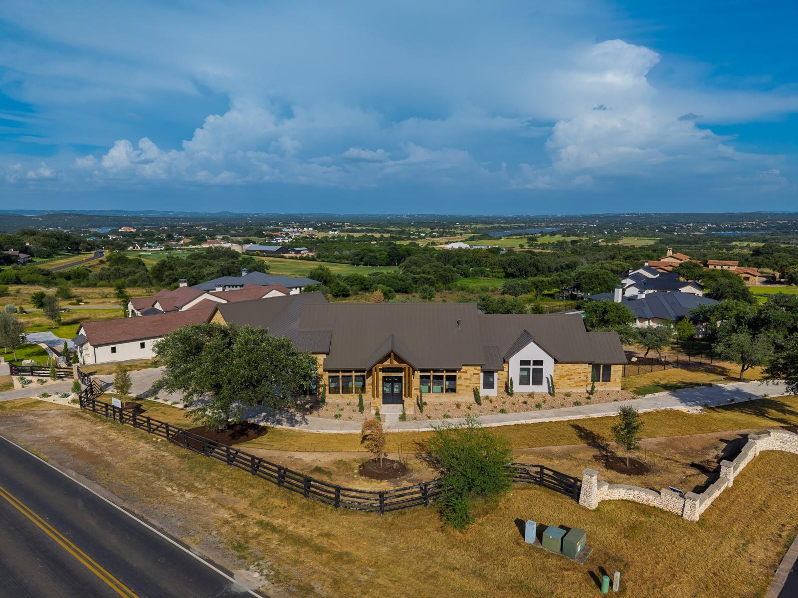 Aerial view of a stone-and-stucco home with a dark roof, fenced yard, and distant lake.