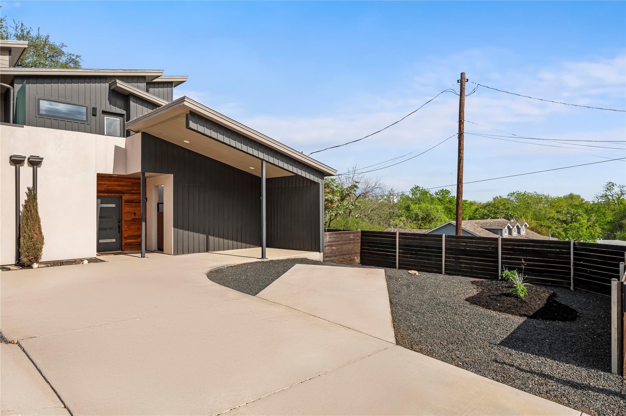 Modern home with dark siding, wood-accented entry, broad sloping roof, and gravel yard behind a black fence.