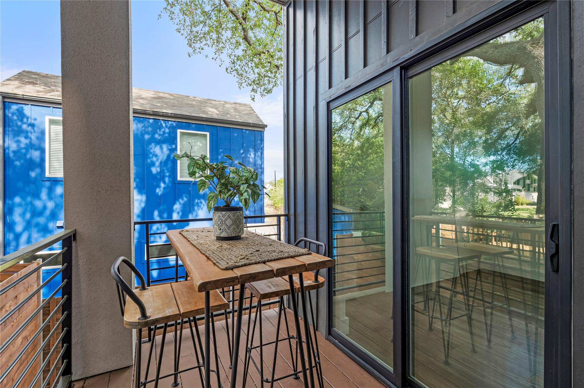 Furnished balcony with a wooden table, potted plant, and sliding glass doors overlooking trees.