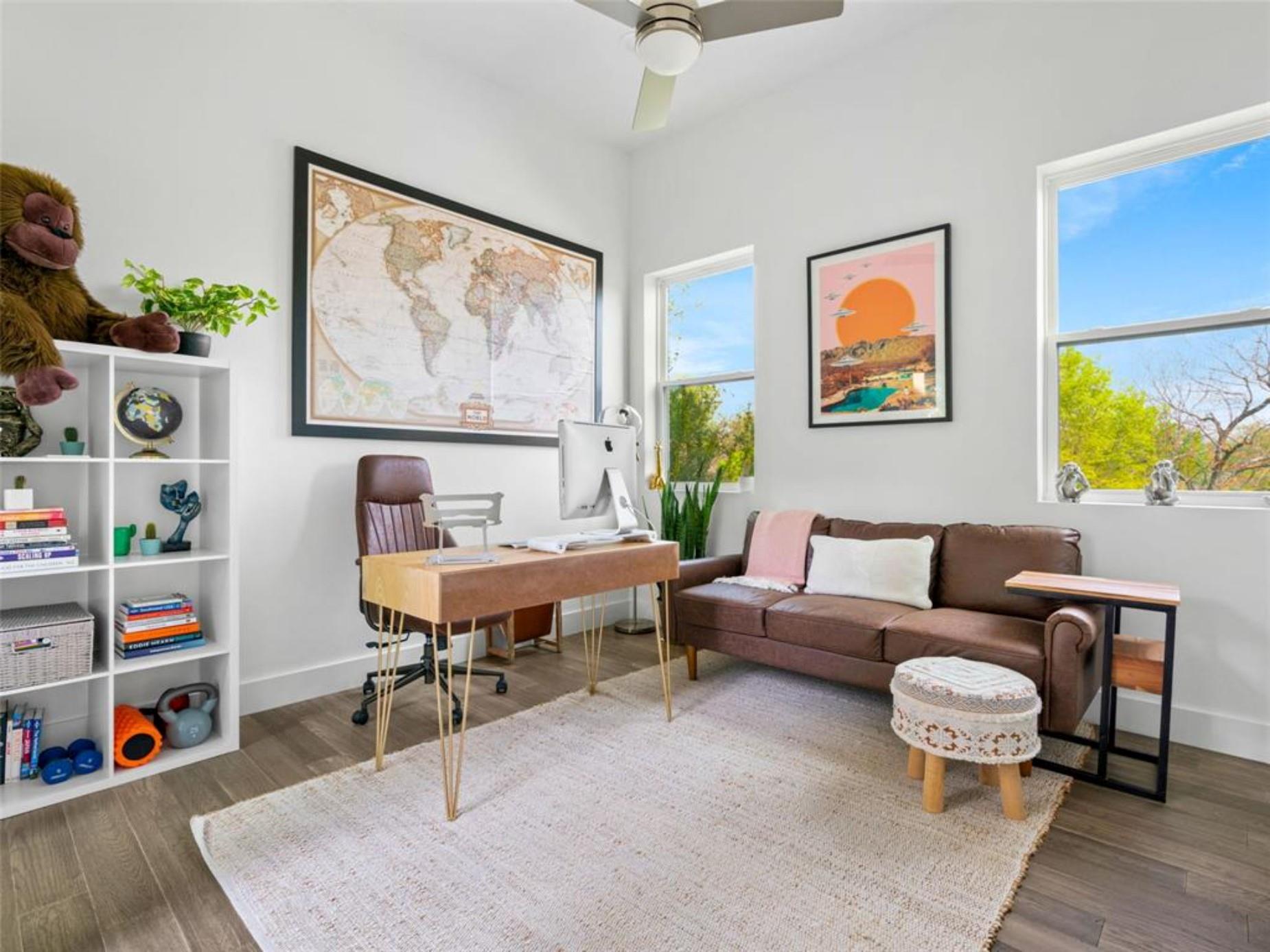 Furnished home office with a wooden desk, brown sofa, white bookcase, and large windows.