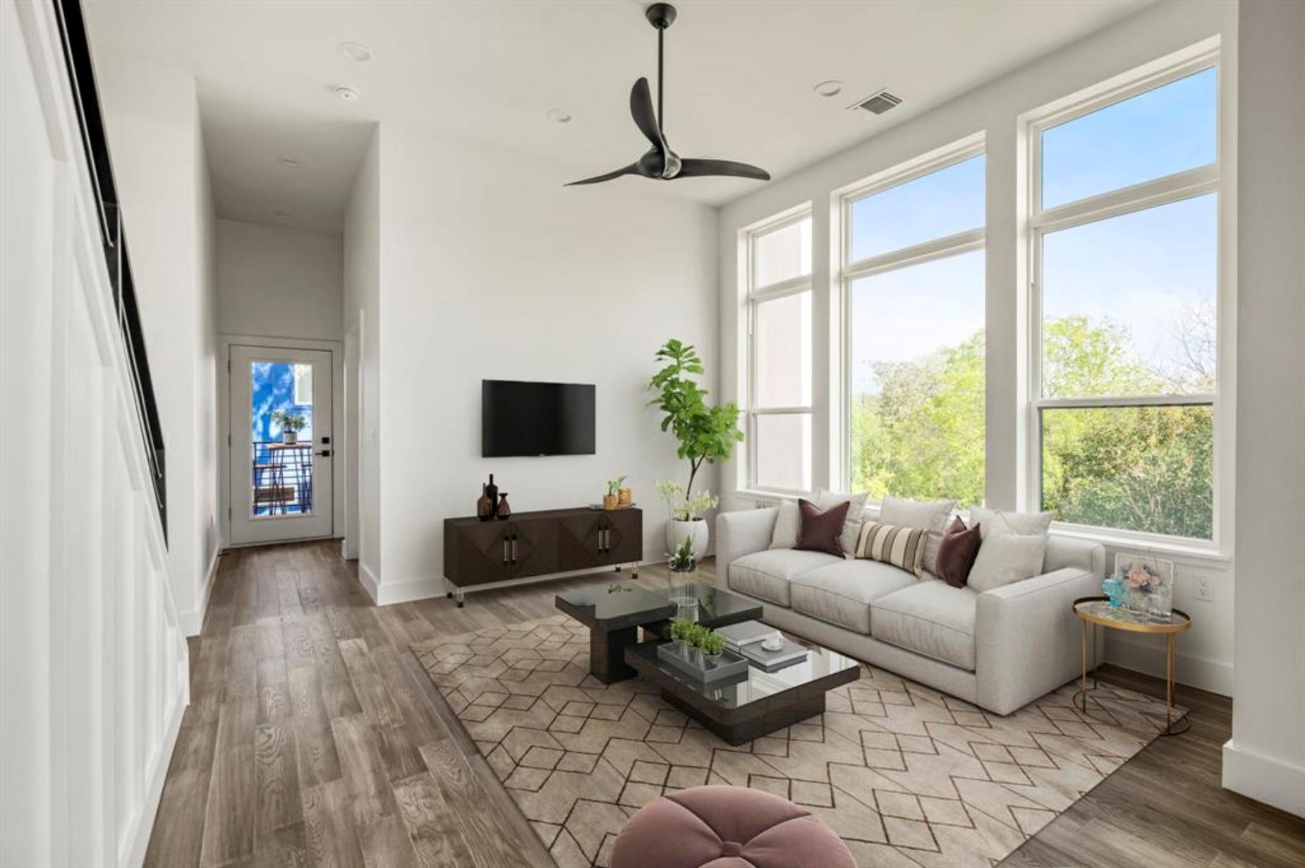 Staged living room with a white sofa, tall windows overlooking trees, and a patterned rug.