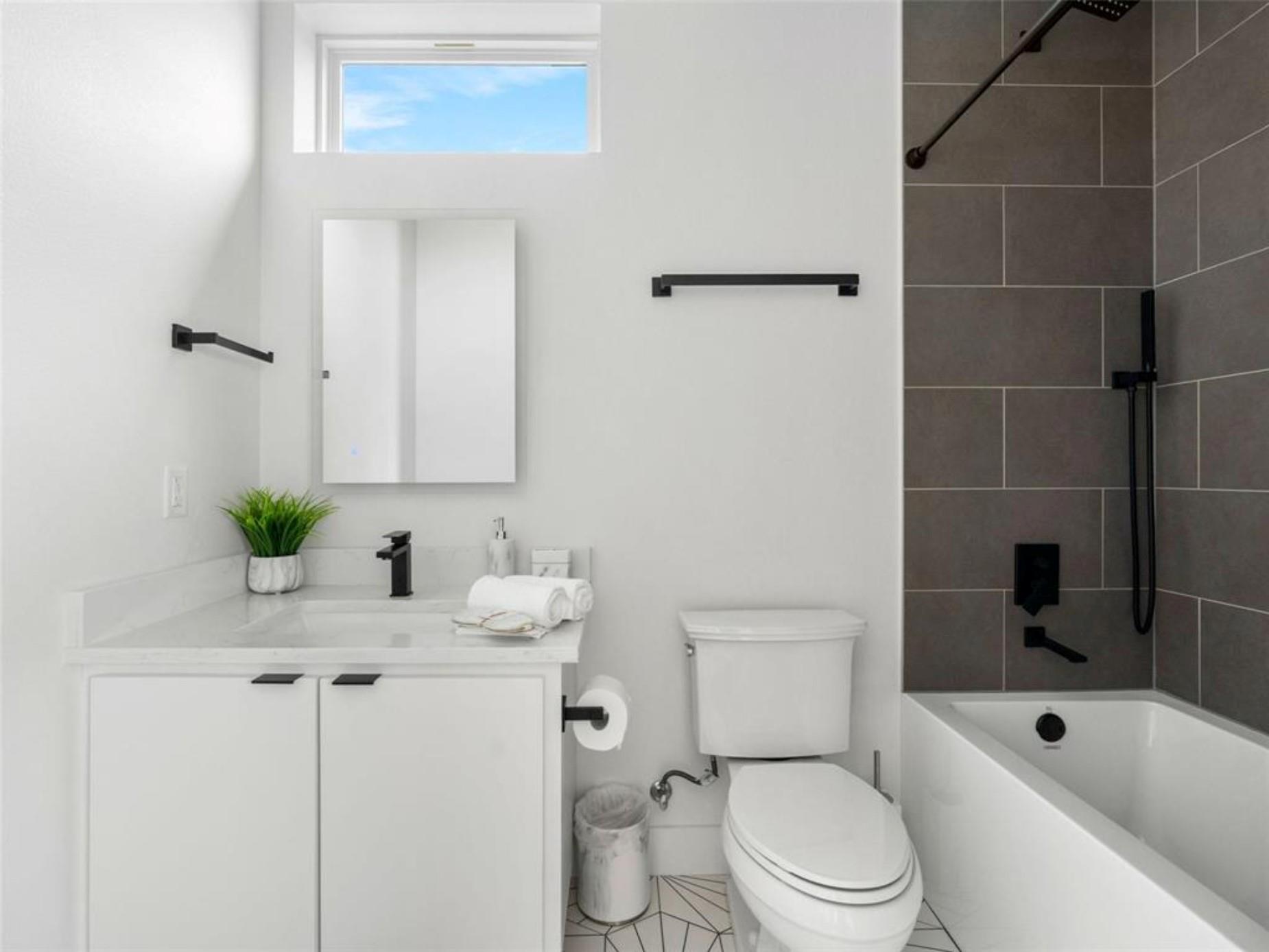 Bright bathroom with a white vanity, toilet, and bathtub beside a dark tiled shower.