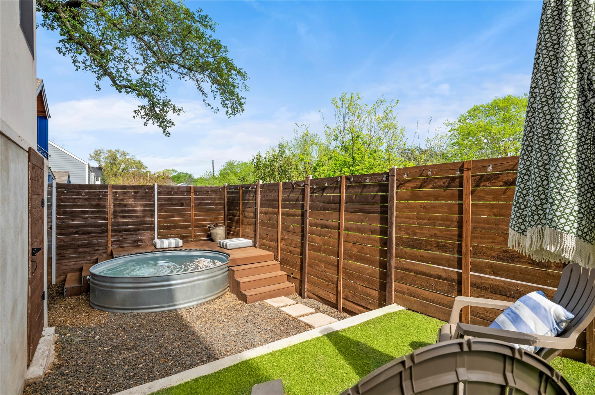 Fenced backyard with a round stock-tank pool, wooden steps, artificial turf, and umbrella-shaded seating.