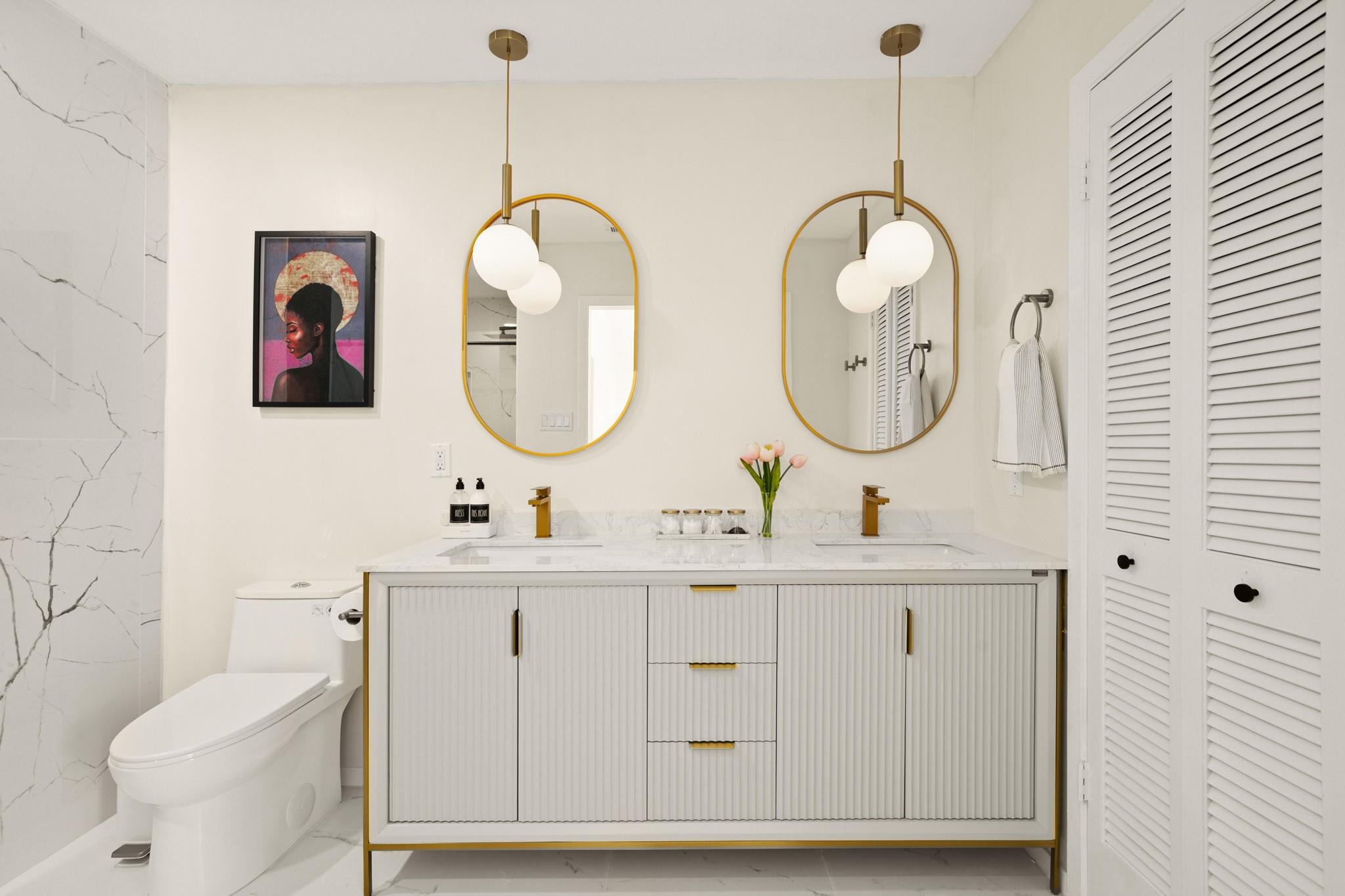 Bright bathroom with a fluted double vanity, brass fixtures, twin oval mirrors, and marble surfaces.