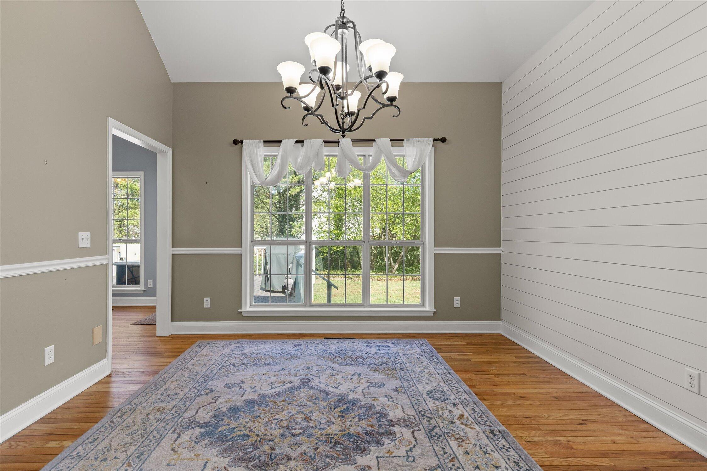 Dining room with a large curtained window, hardwood floors, patterned rug, and white shiplap wall.