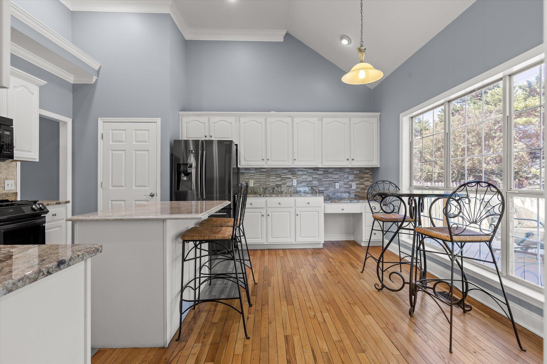 Kitchen with white cabinets, stainless appliances, hardwood floors, and a breakfast table by large windows.