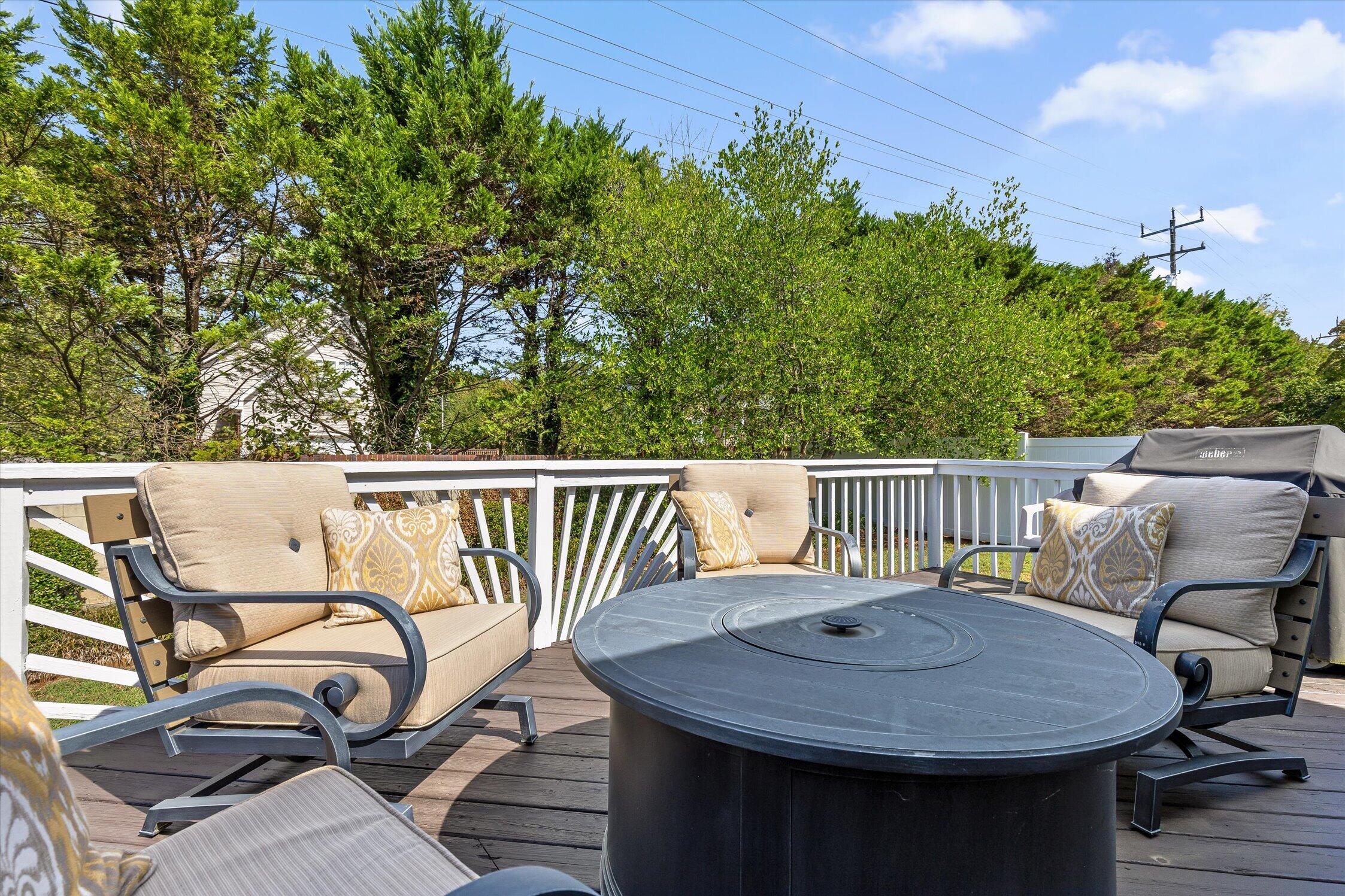 Furnished deck with cushioned chairs around a round black fire table and white railings.