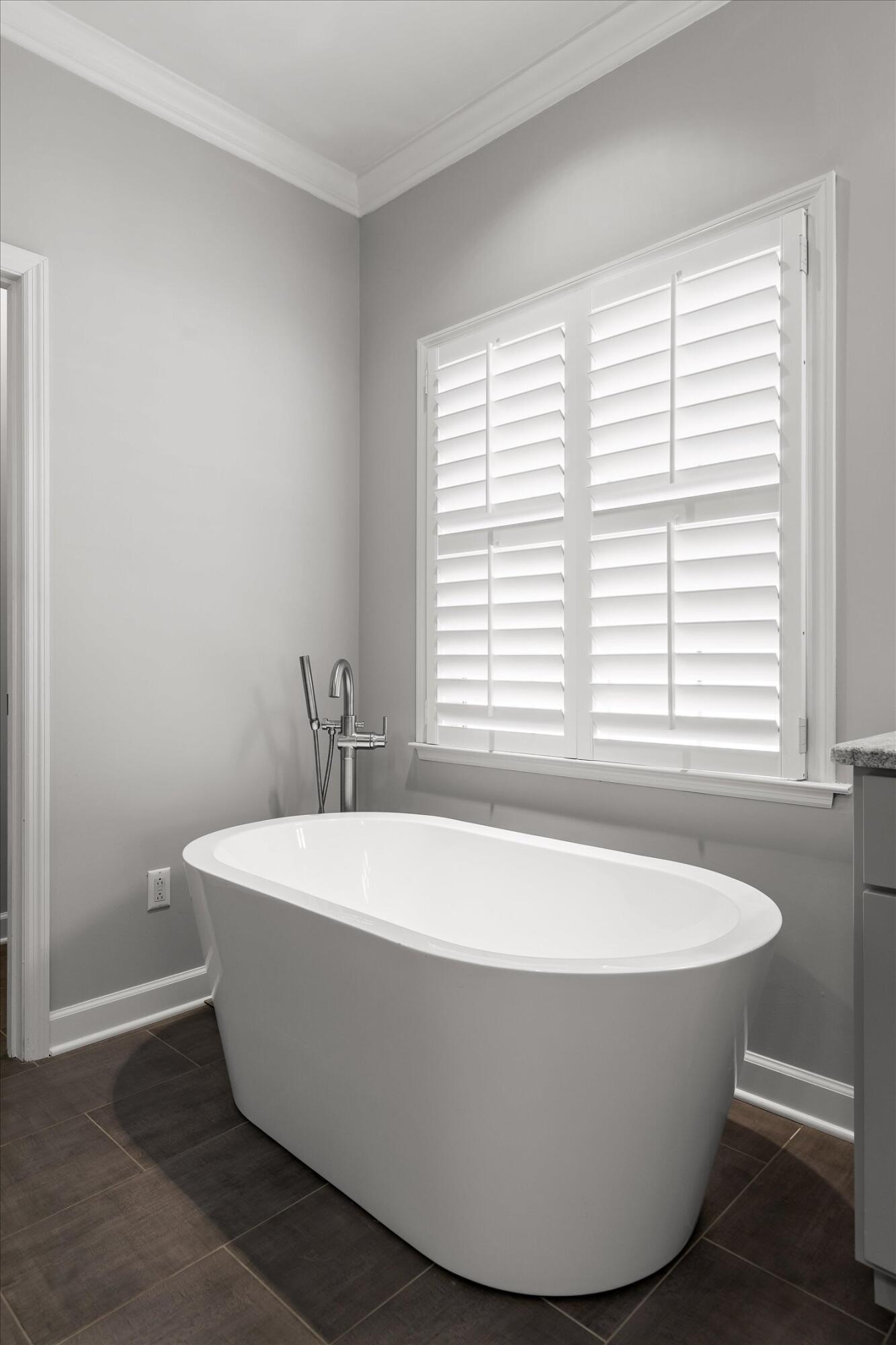 Freestanding white soaking tub beneath a large window with plantation shutters.