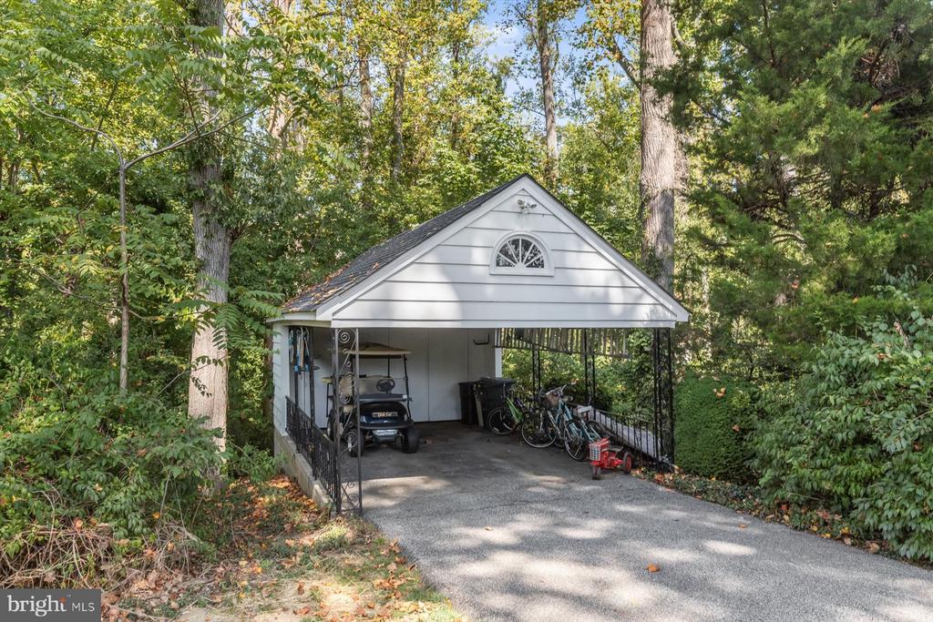 Detached white carport with a peaked roof, golf cart, and bicycles amid mature trees.