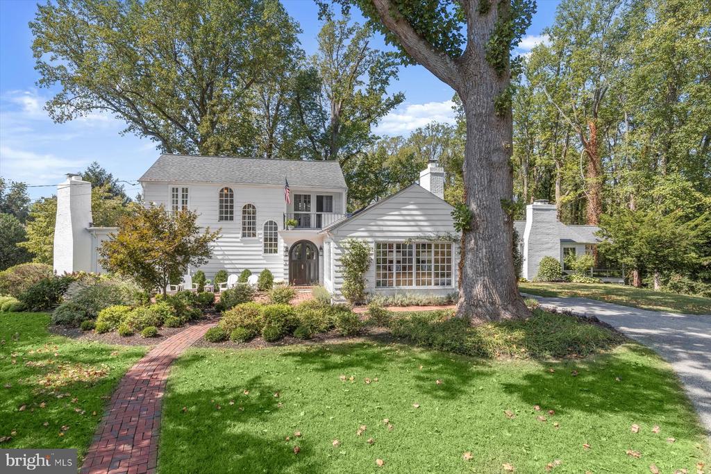 White two-story house behind a large mature tree, with a brick walkway and landscaped front lawn.