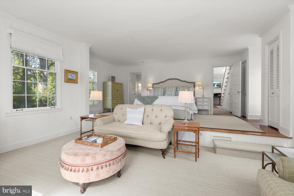 Bedroom with a raised bed, cream sofa, pink ottoman, and large windows overlooking trees.