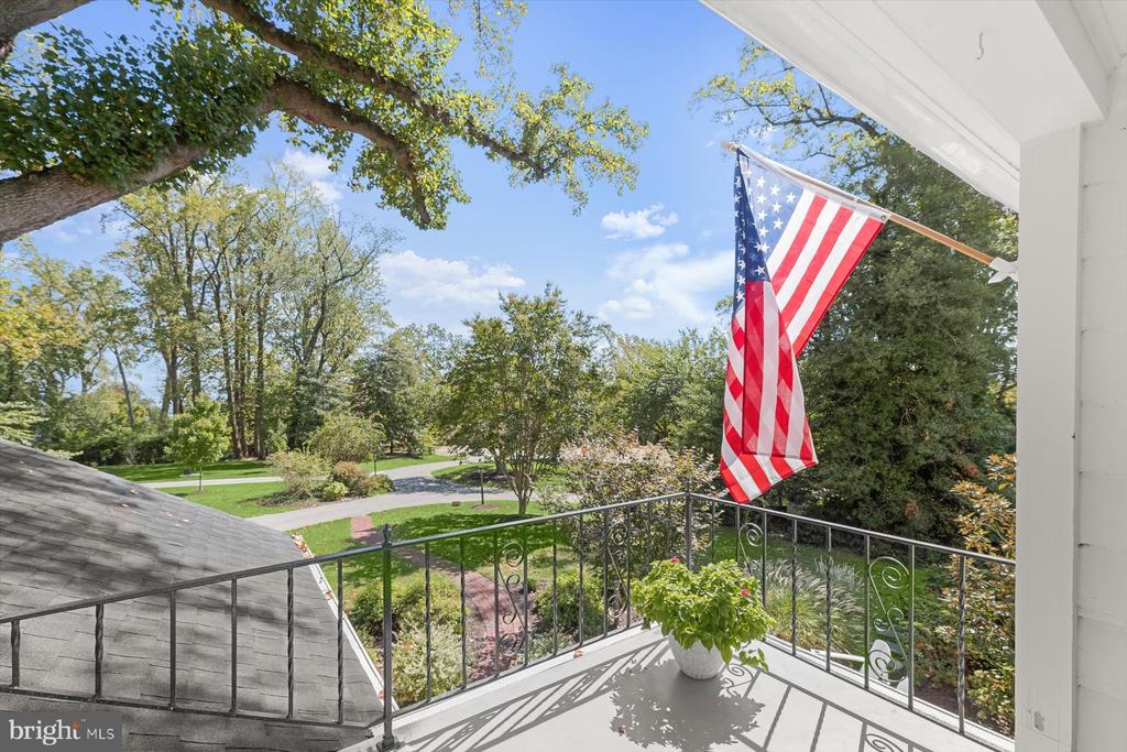 Covered balcony with an American flag overlooking landscaped grounds and a tree-lined road.