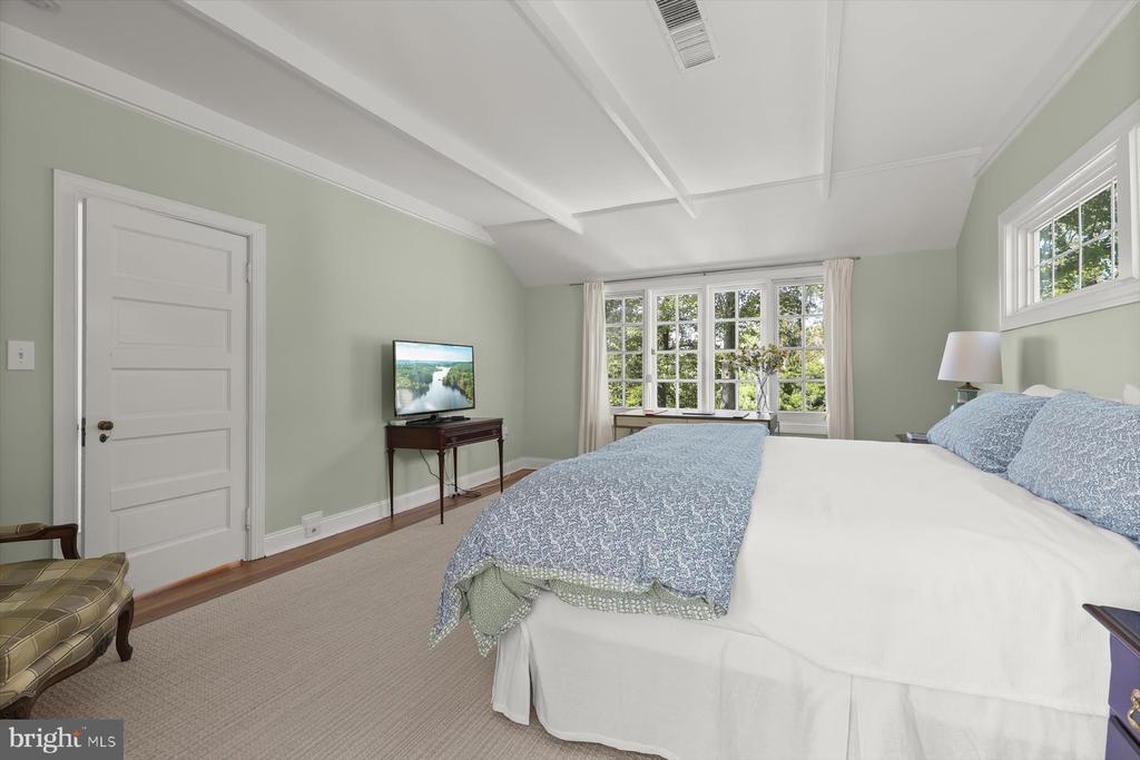 Bedroom with blue patterned bedding, white ceiling beams, and a wide window overlooking trees.
