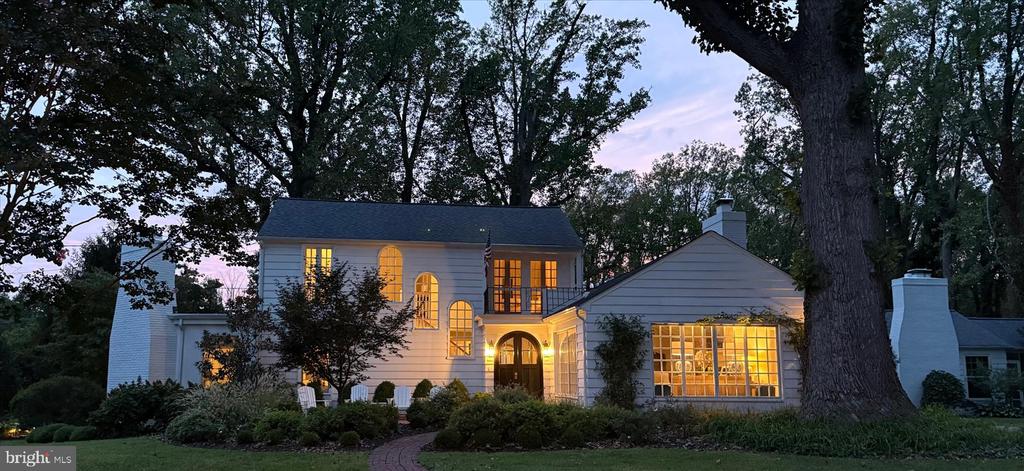 White house at dusk with warmly lit windows, an arched entry, and mature trees.