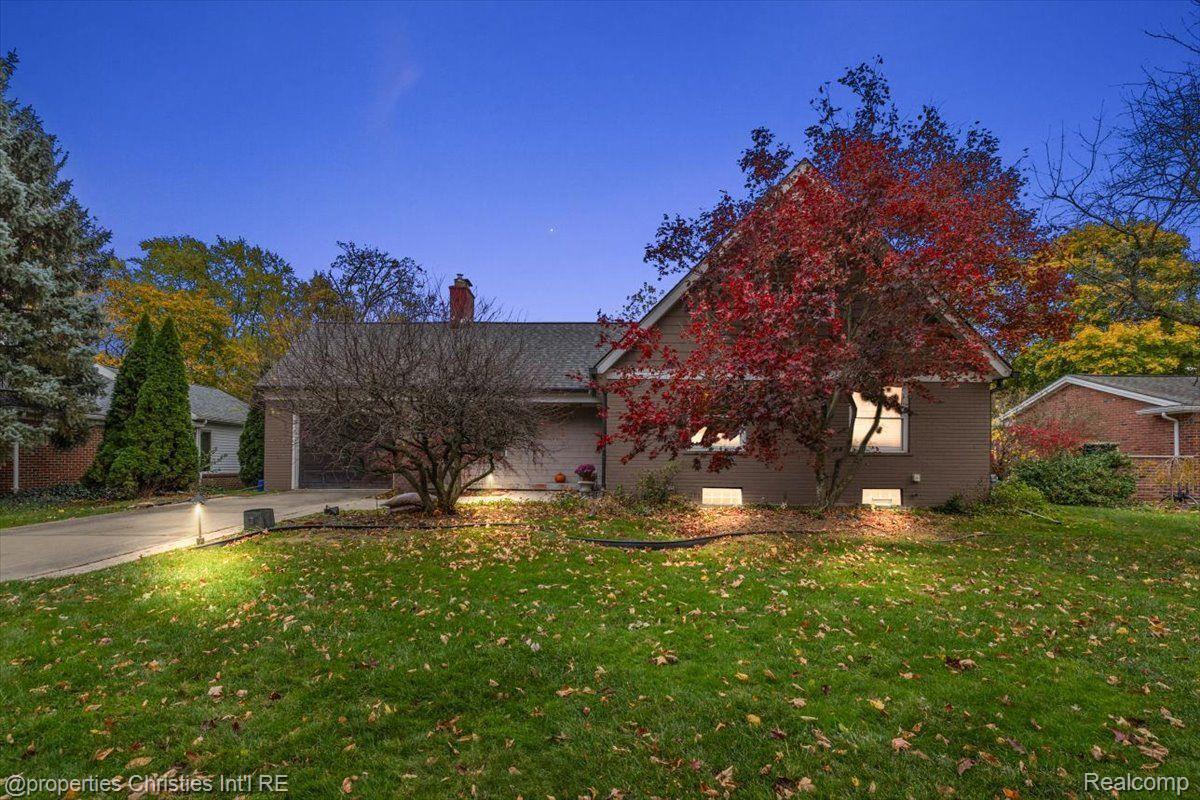 Single-story brown house at dusk, with a red autumn tree, broad driveway, and leaf-strewn lawn.
