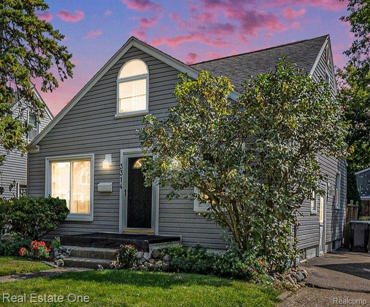 Gray two-story house with an arched dormer, front steps, and leafy landscaping at dusk.