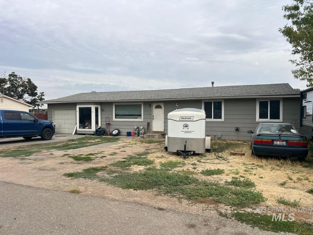 Single-story gray house with an attached garage, white enclosed trailer, and vehicles parked in front.