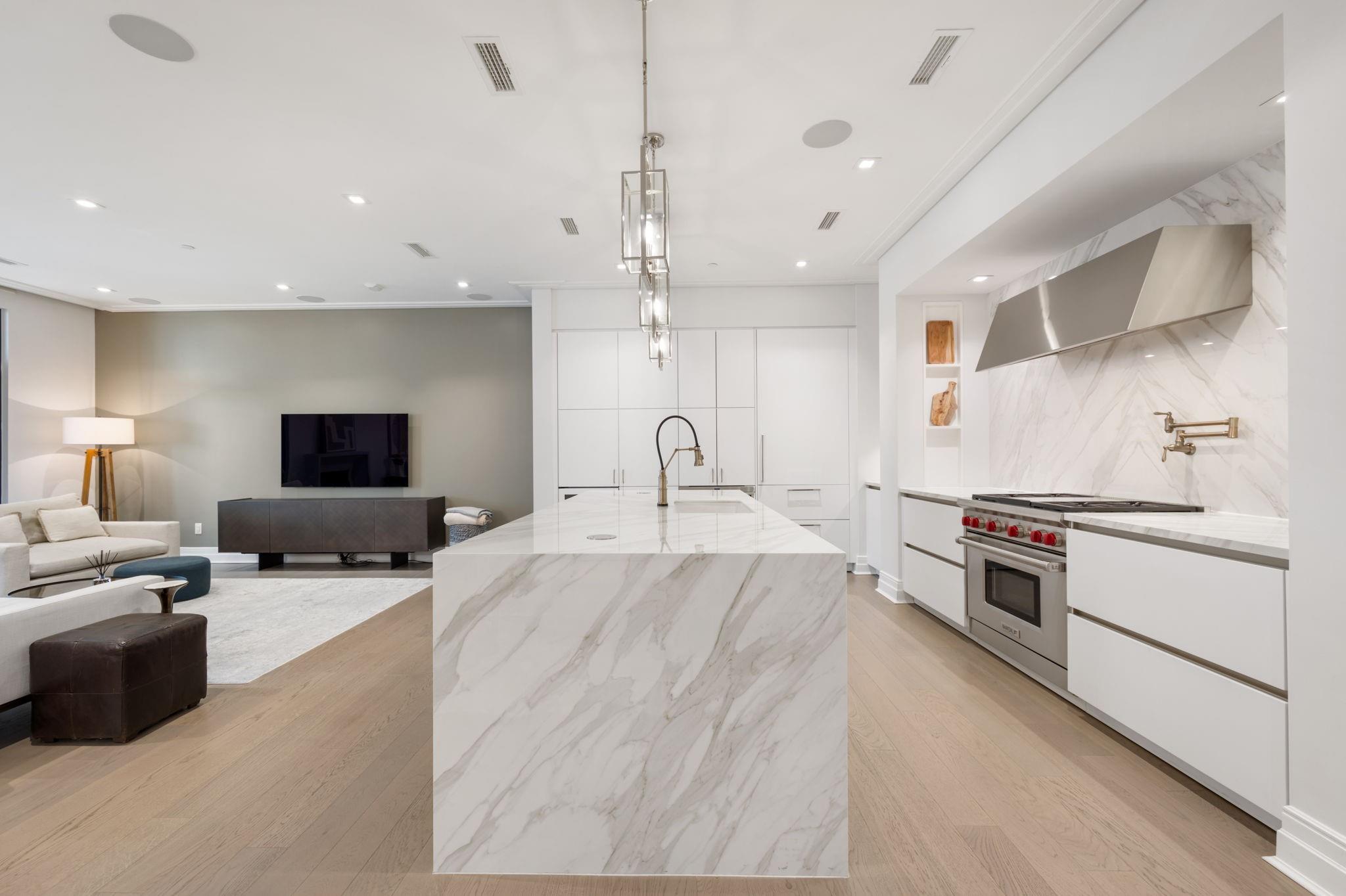 Modern kitchen with a large marble island, stainless range, and adjoining furnished living area.