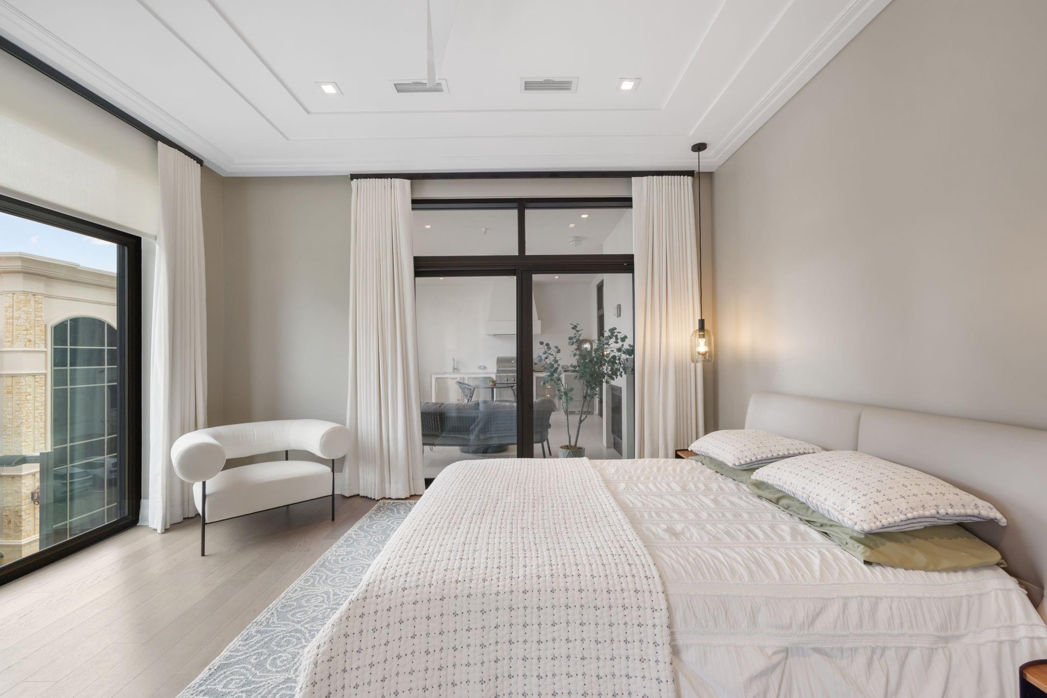 Bright bedroom with a large white bed, sculptural chair, and black-framed glass doors.
