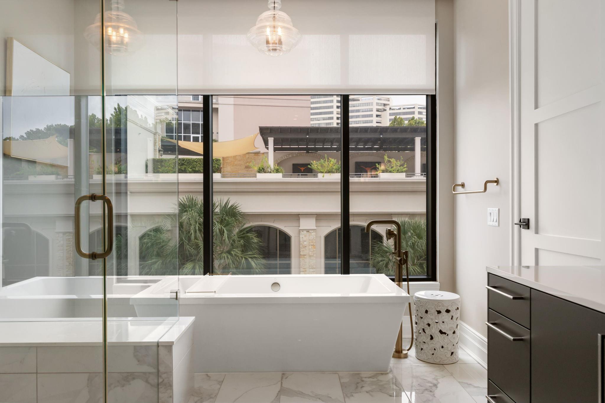 Modern bathroom with a freestanding soaking tub, glass shower, marble floor, and large courtyard-facing windows.