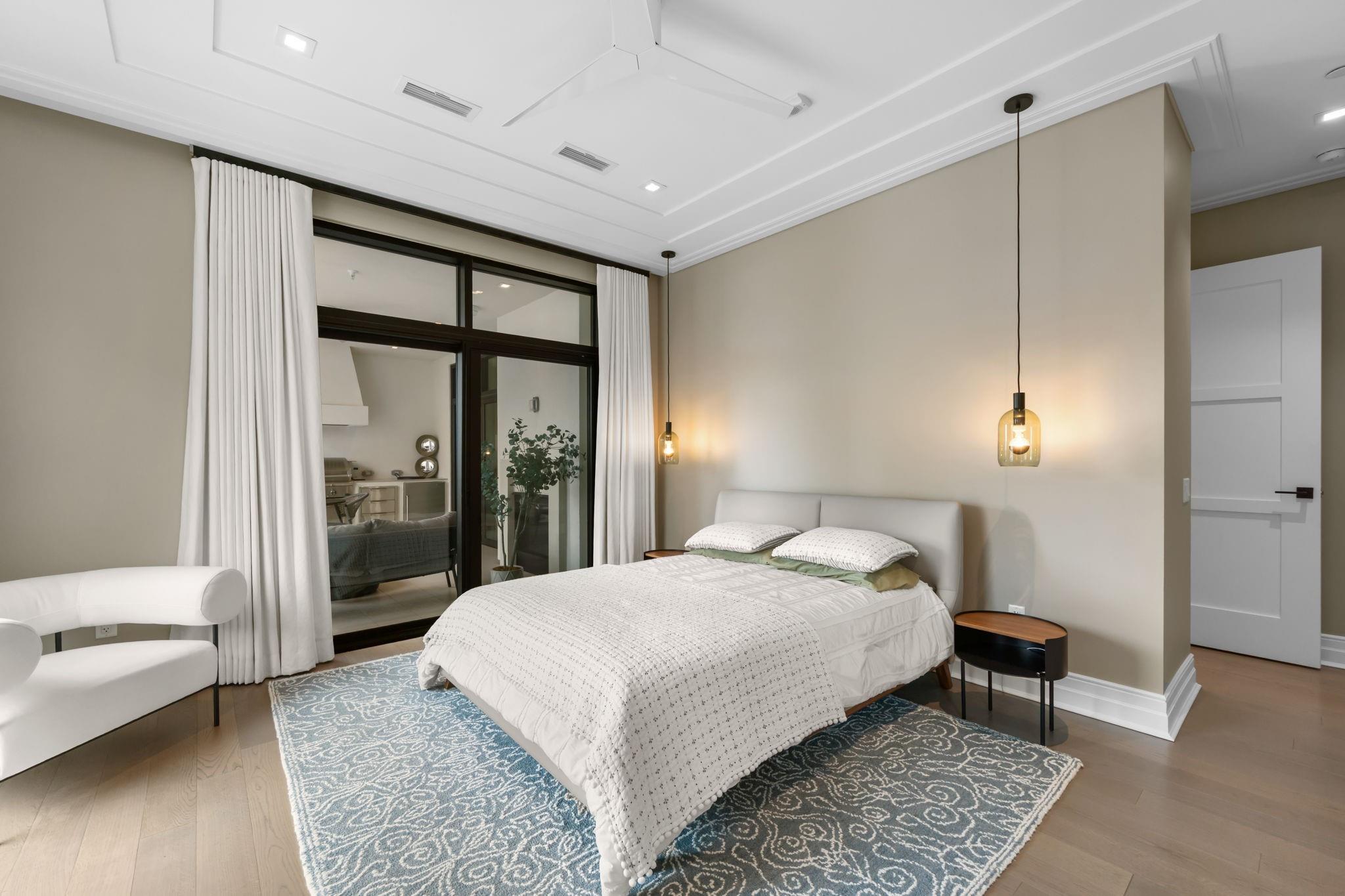 Staged bedroom with a white bed, patterned rug, and glass doors framed by white curtains.