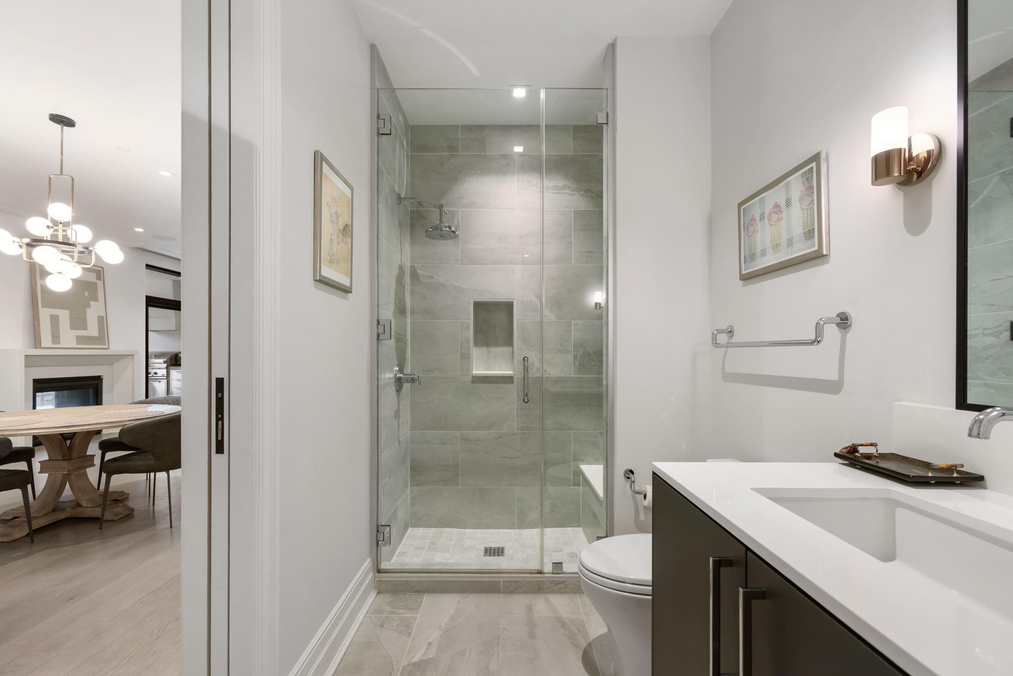 Bathroom with a glass-enclosed gray-tile shower and white vanity, opening onto a dining area.